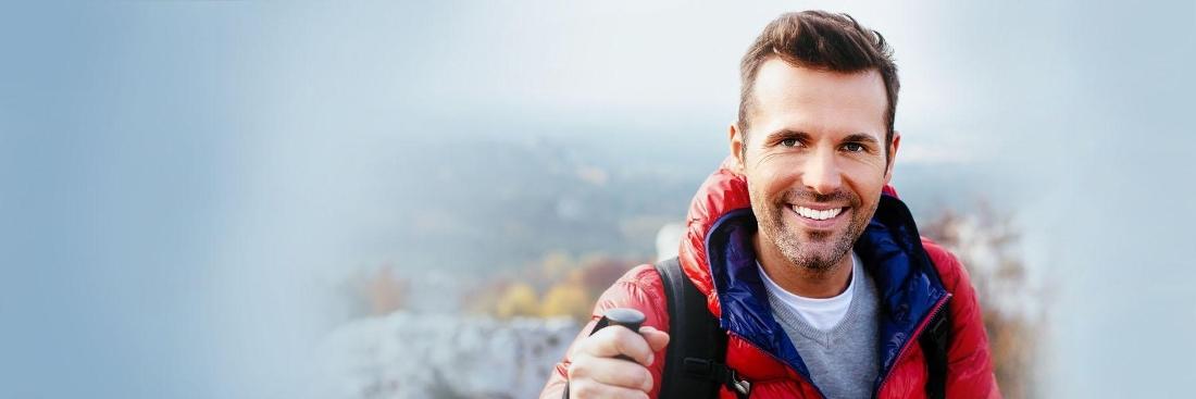 brown haired man holding a hiking stick and smiling against a mountainous background